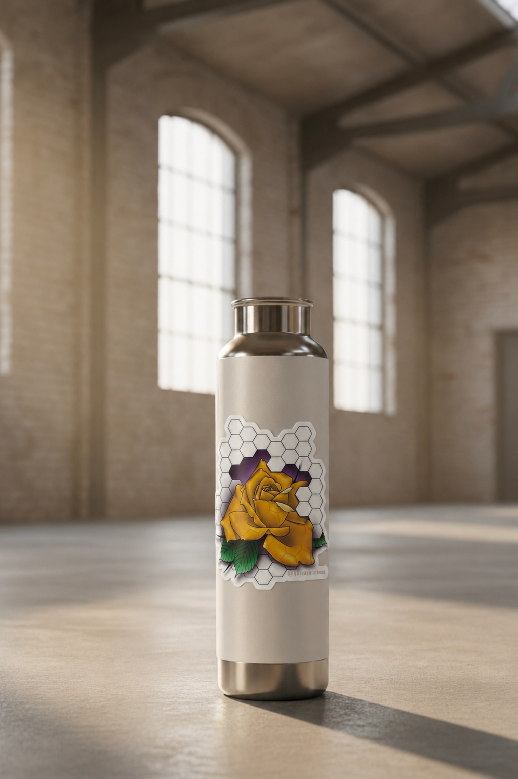 Metal water bottle with a gold rose design on a wooden floor in a large room with high ceilings and large windows.